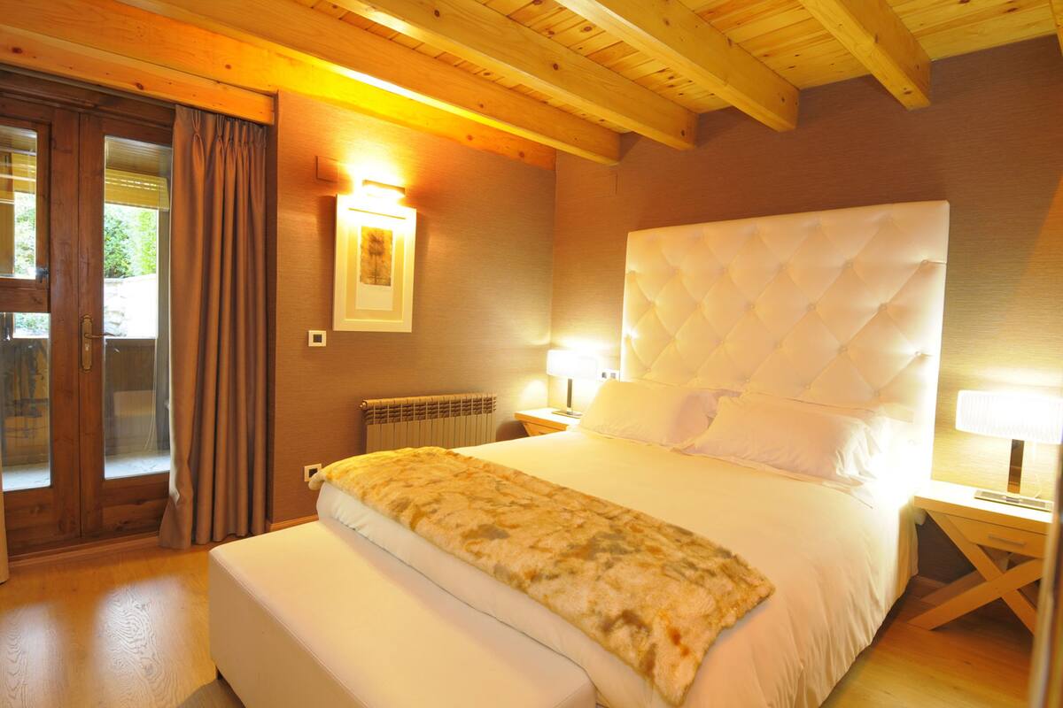 A bedroom with a large bed, wooden ceiling beams, a radiator, and a window with curtains.