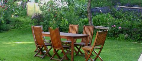 Juego de comedor exterior de madera con cuatro sillas y una mesa, rodeado por un césped verde y diversas plantas.