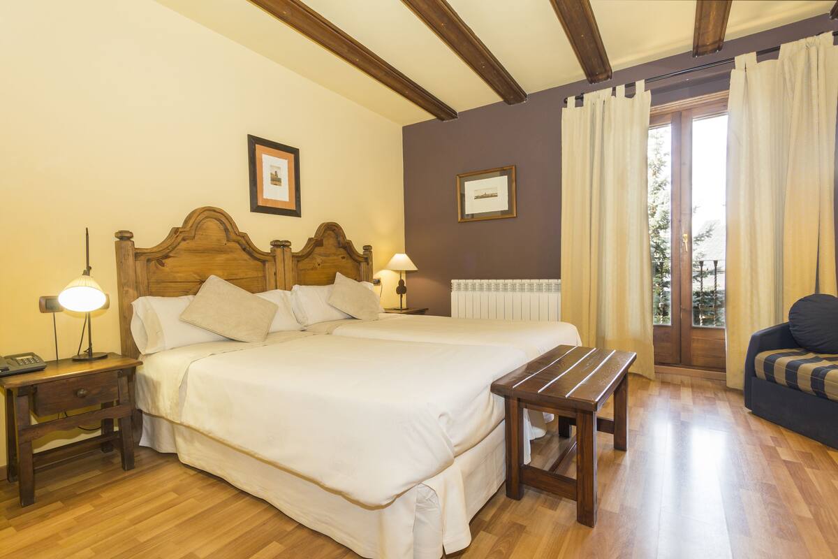 A hotel room with a large bed, bedside tables, a wooden bench, and a view of the outdoors through a window with curtains.