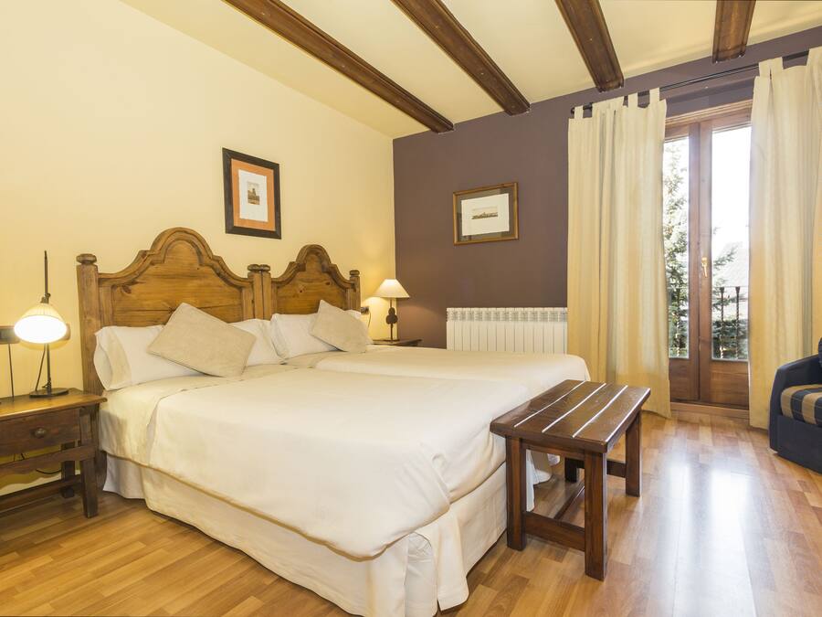 A hotel room with a large bed, bedside tables, a wooden bench, and a view of the outdoors through a window with curtains.