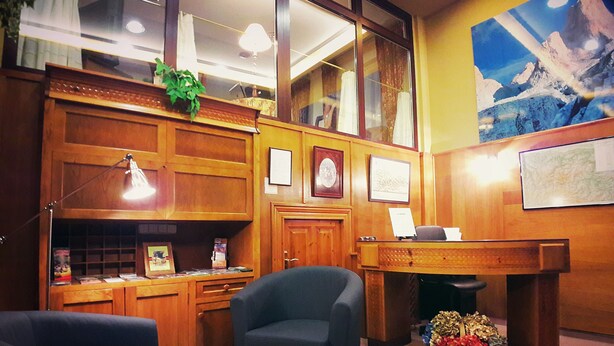 A reception area with a wooden desk, a blue chair, a lamp, framed certificates, and a large window with curtains. - Hotel La Rivera (Cabrales)