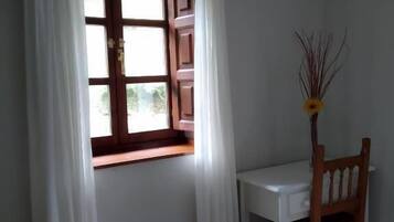 A room with a wooden desk and chair, a window with white curtains, and a vase with a yellow flower.