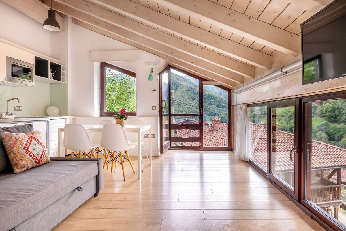 A spacious living area with a wooden ceiling, a sofa, a dining table with chairs, and large windows offering a view of a mountainous landscape.