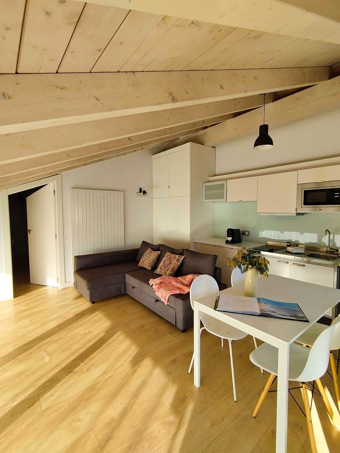 Una cocina moderna con armarios blancos y un microondas, un comedor con mesa y sillas blancas, y una sala de estar acogedora con un sofá gris y una manta rosa.