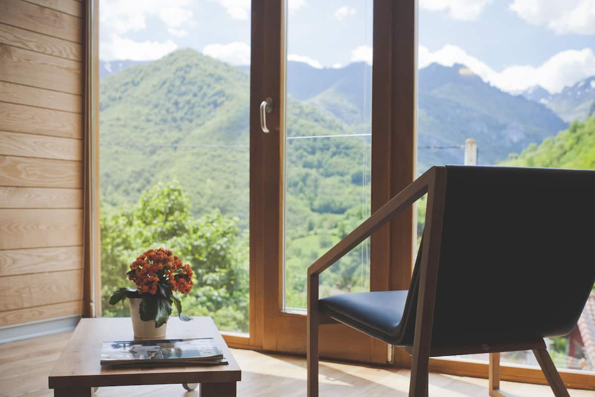 A room with a wooden floor, a table with a vase of flowers, a chair, and a large window offering a mountain view.