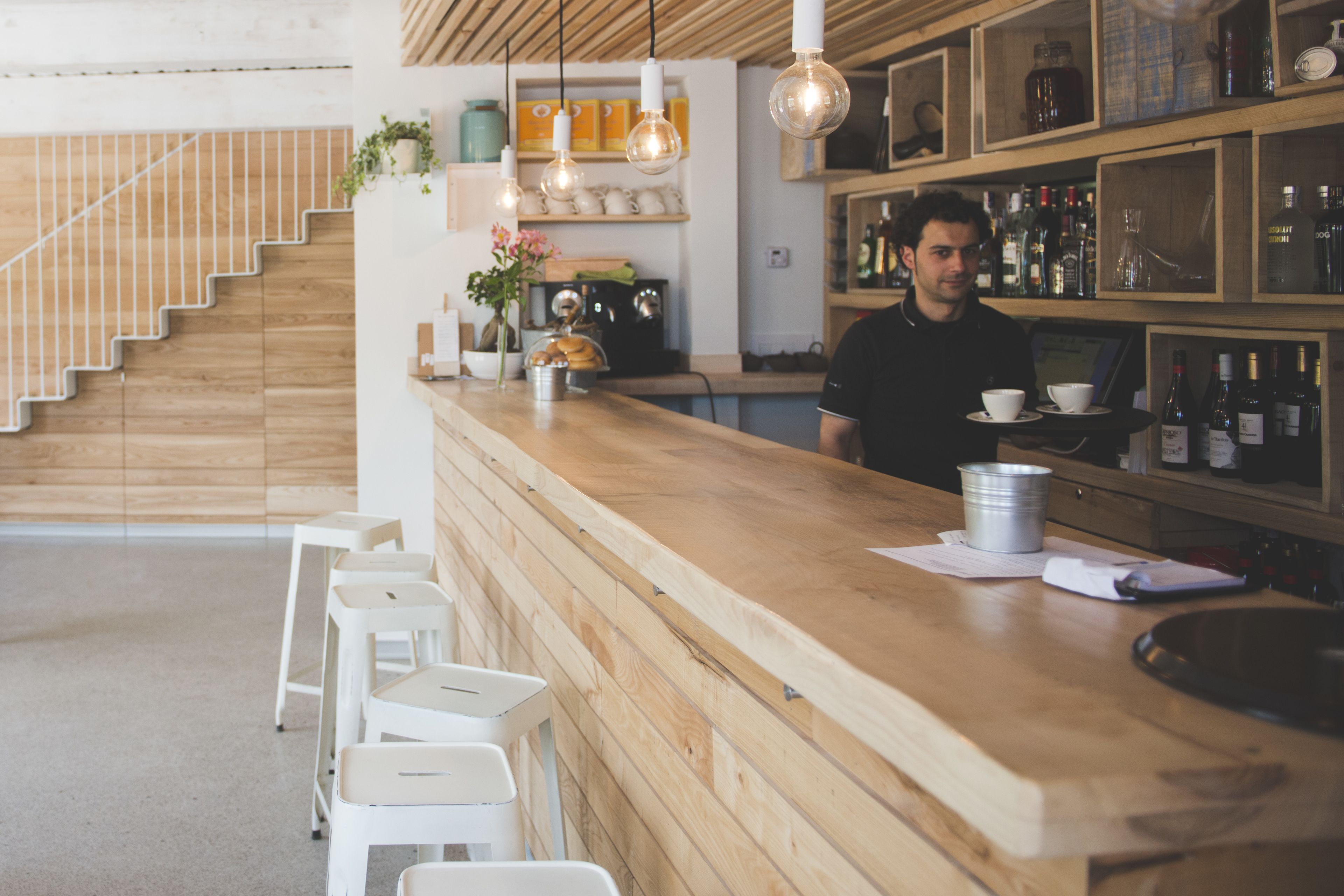 Una zona de bar con mostrador de madera, taburetes blancos, lámparas colgantes y estantes con botellas.