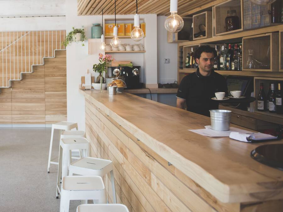 Una zona de bar con mostrador de madera, taburetes blancos, lámparas colgantes y estantes con botellas.