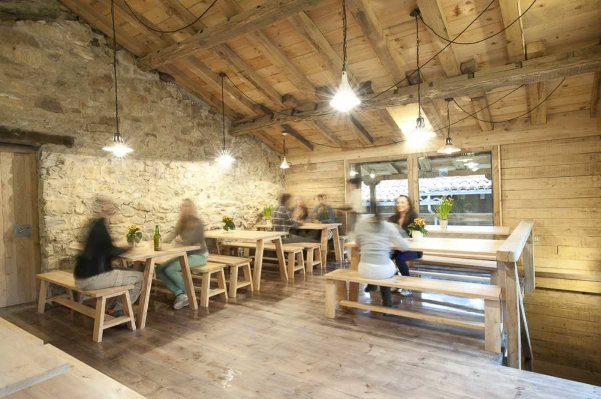 A rustic wooden interior with exposed beams and stone walls, featuring wooden tables and benches arranged for dining.