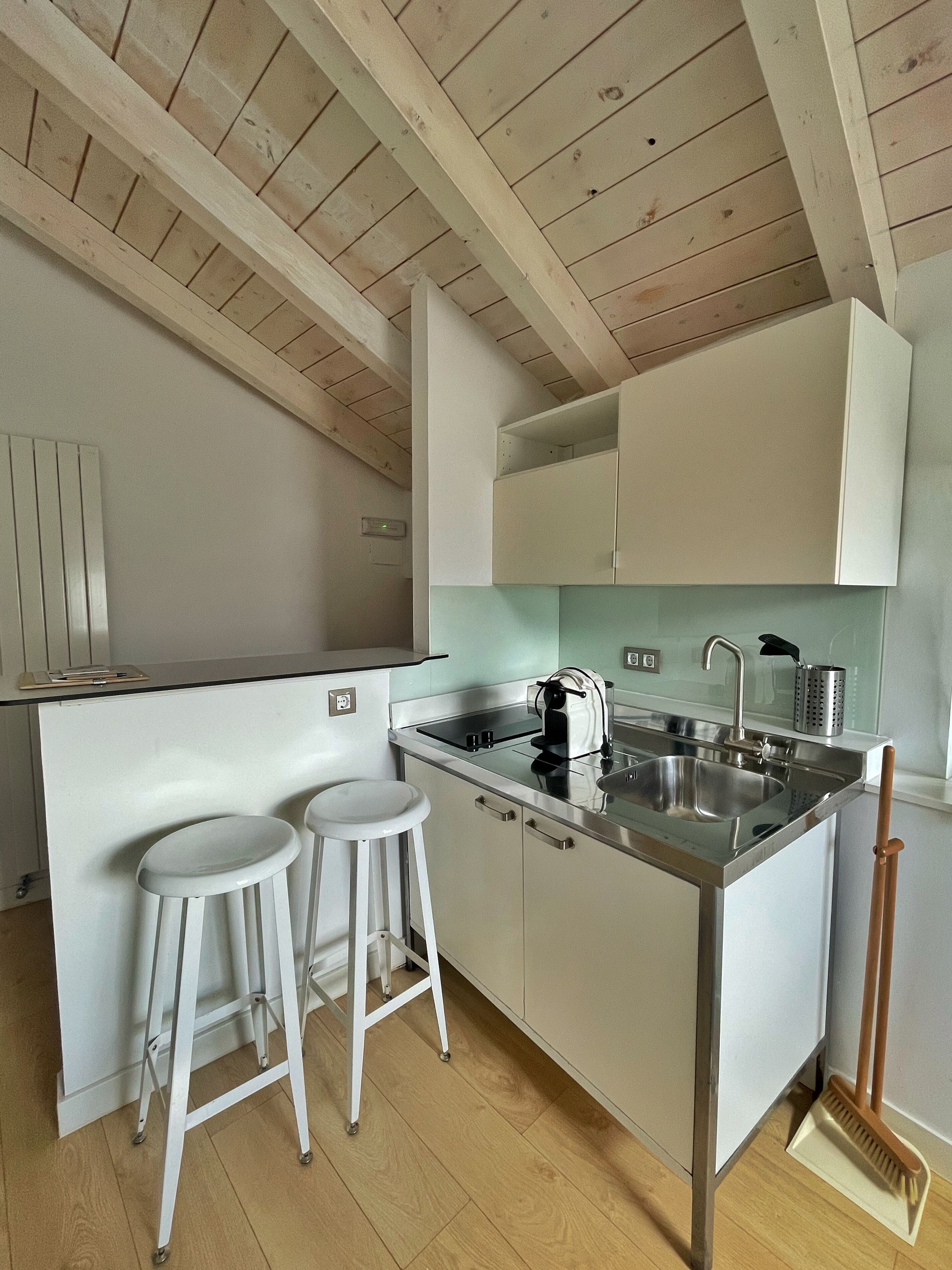 A compact kitchen with white cabinets, a stainless steel sink, two stools, and a wooden ceiling.
