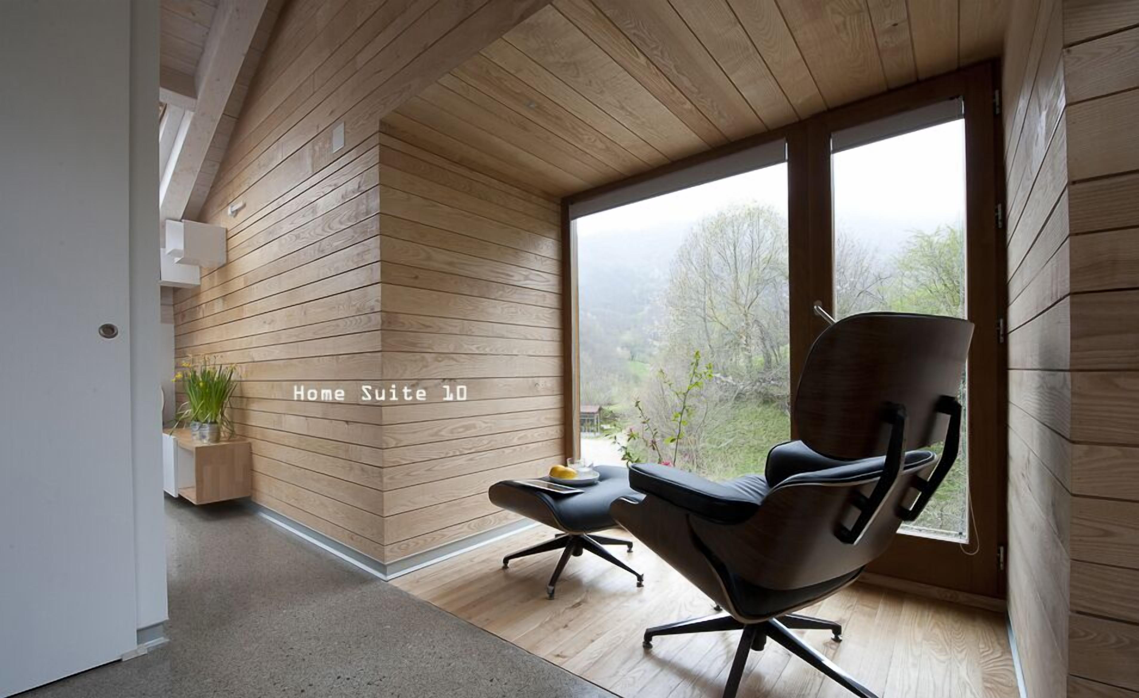 A modern interior with wooden panel walls, a black office chair, a small table with a fruit bowl, and a view of a forest through large windows.