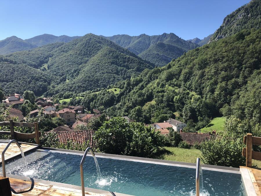 A pool with clear water and a wooden deck, surrounded by lush green mountains and a small village.