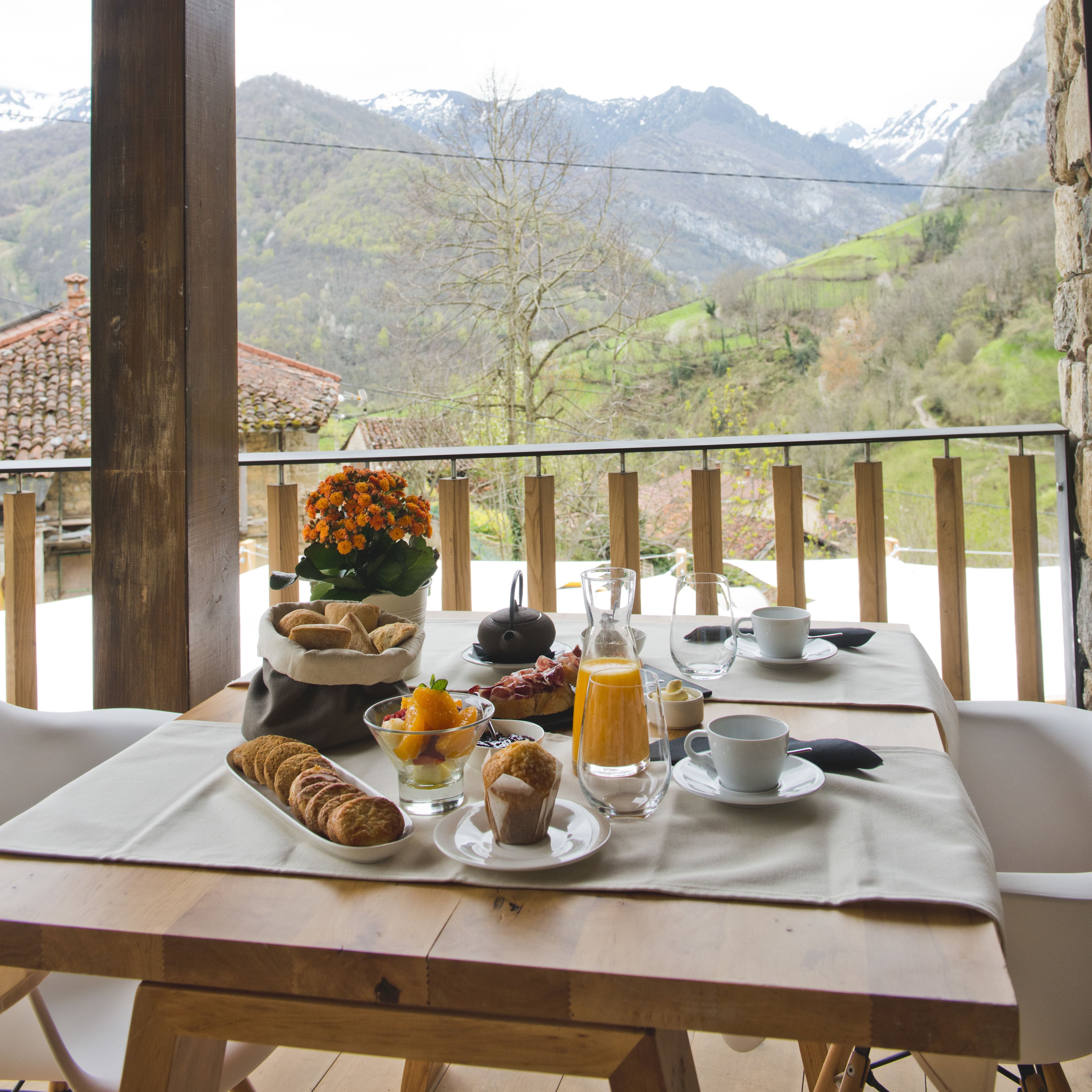 Une table dressée pour le petit-déjeuner, avec des viennoiseries, des fruits et des boissons, donnant sur un paysage montagneux.