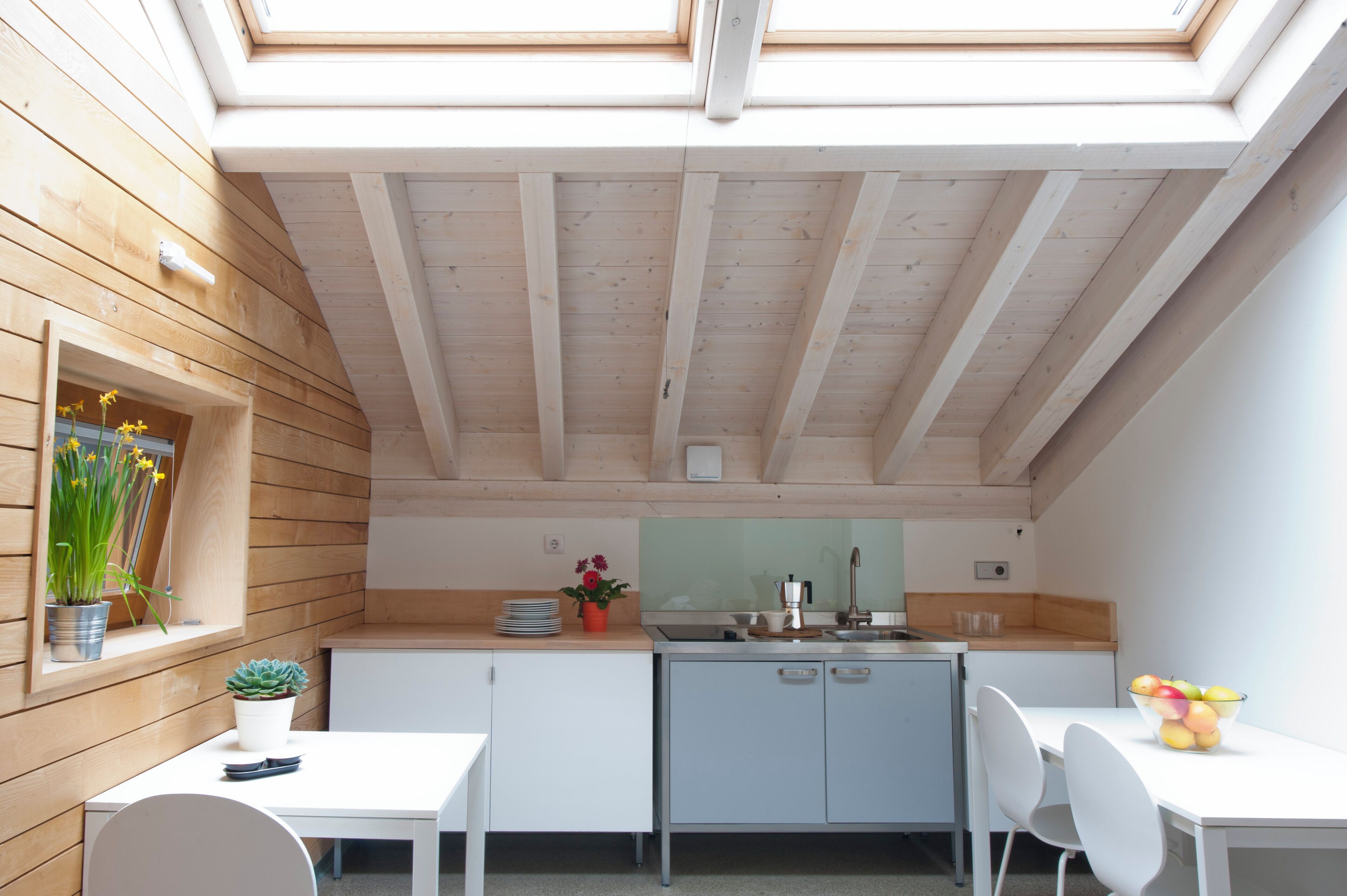 Cocina compacta con vigas de madera, armarios blancos, tragaluces y un comedor con sillas.