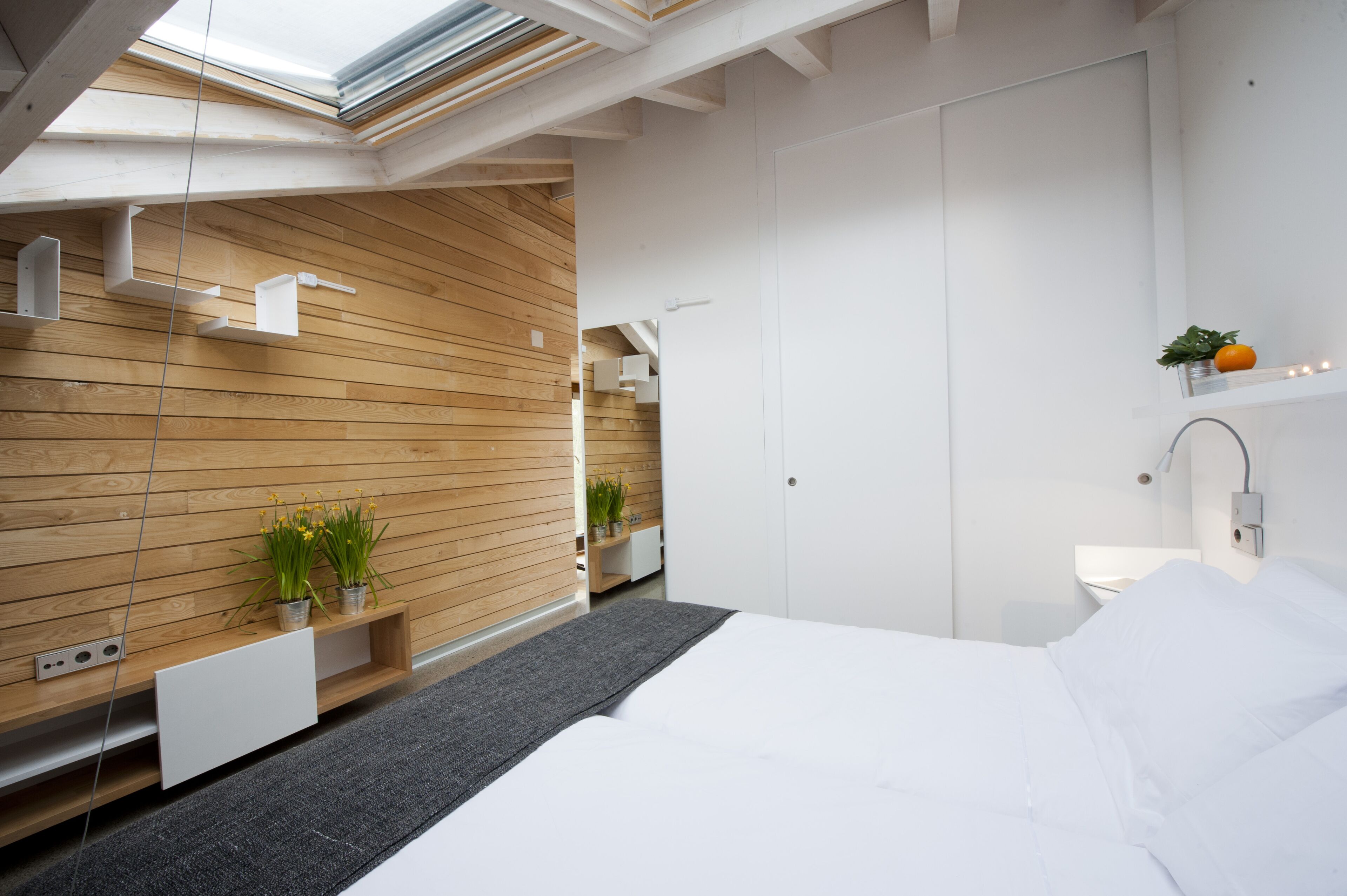 Un dormitorio moderno con tragaluz, paneles de madera, una cama blanca y una planta pequeña en la mesita de noche.
