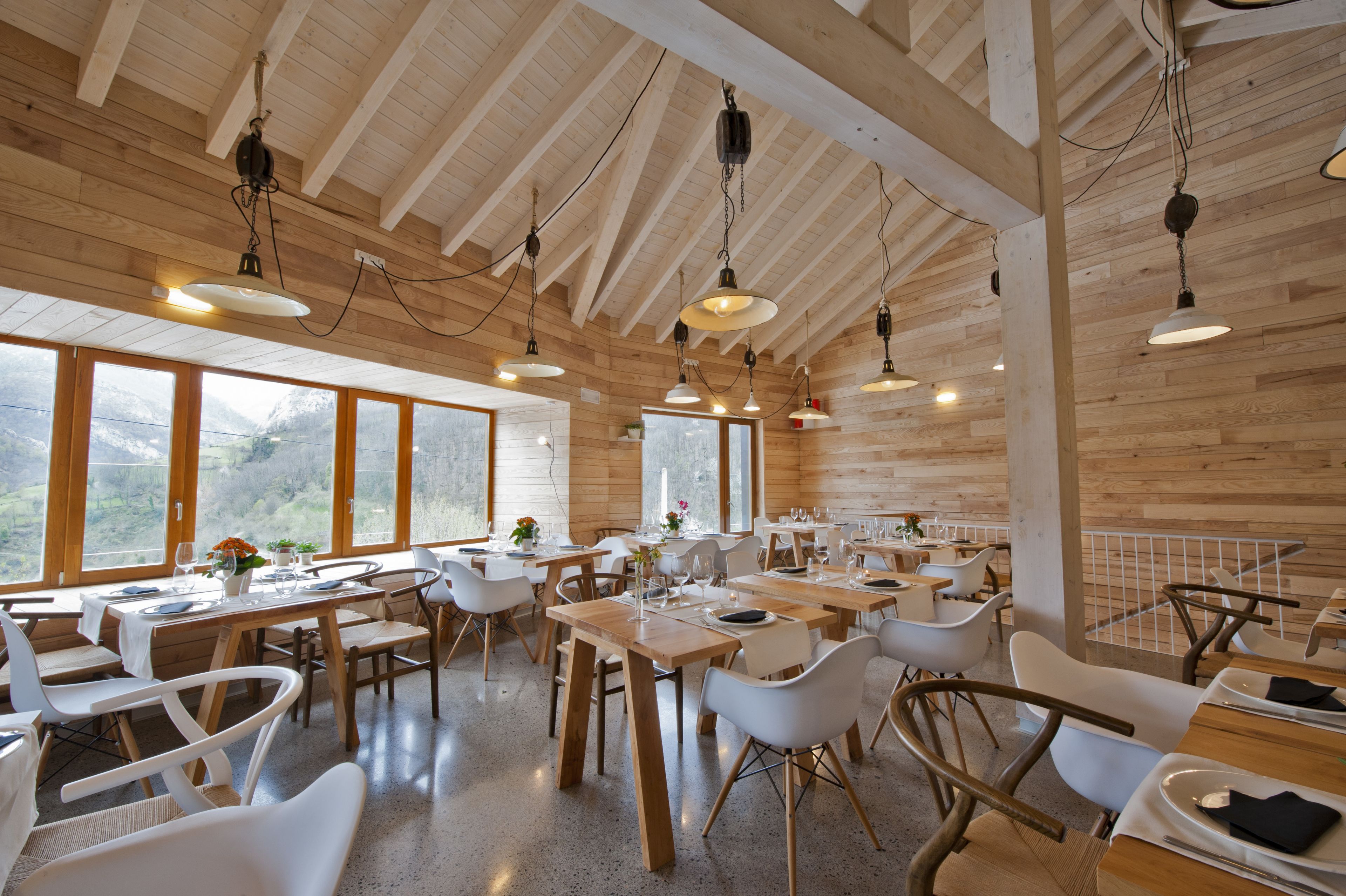 A spacious dining area with wooden tables and white chairs, large windows offering mountain views, and hanging pendant lights.