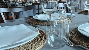 Una mesa de comedor dispuesta con platos, cubiertos y cristalería transparente, con individuales tejidos y vistas al exterior a través de grandes ventanales.
