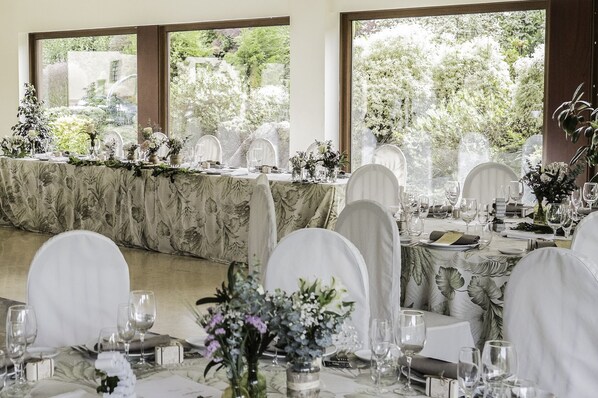 A banquet hall with a long table set for a formal event, surrounded by chairs with white covers, and large windows offering a view of greenery outside.