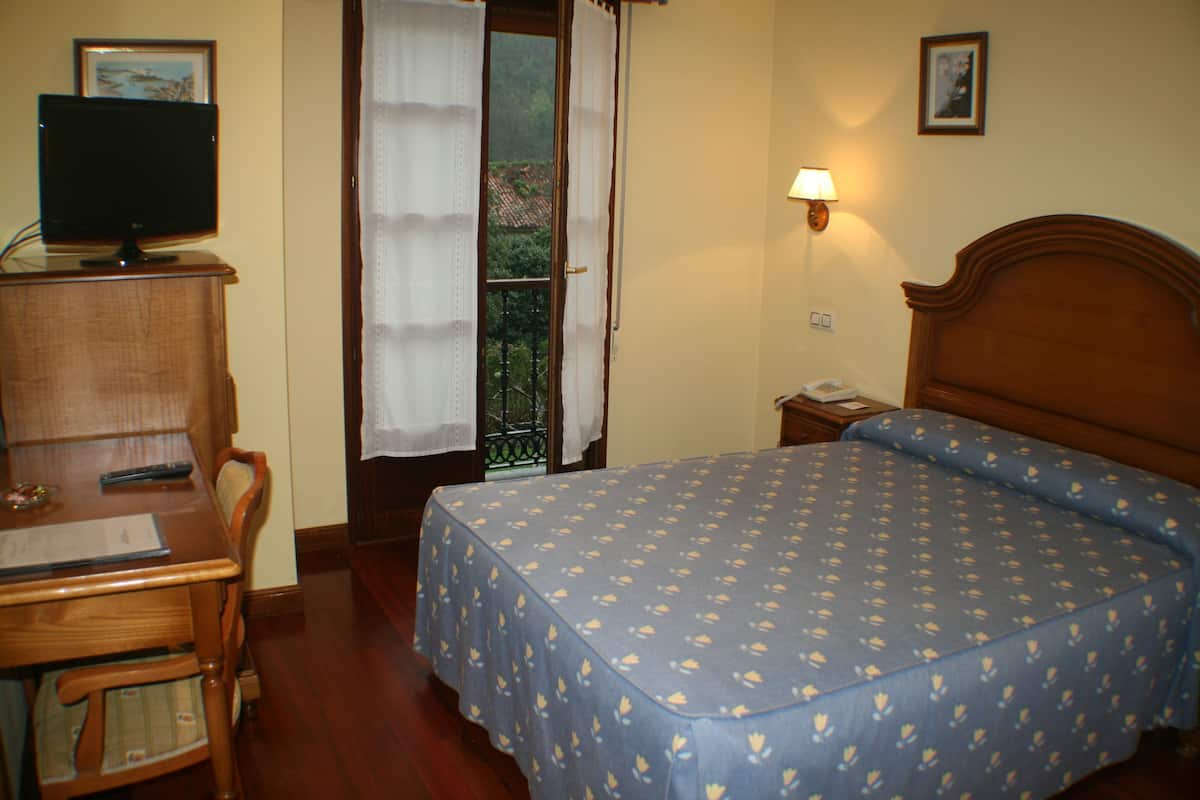 A hotel room with a bed, a desk with a computer, a chair, a nightstand with a lamp, and a window with curtains.