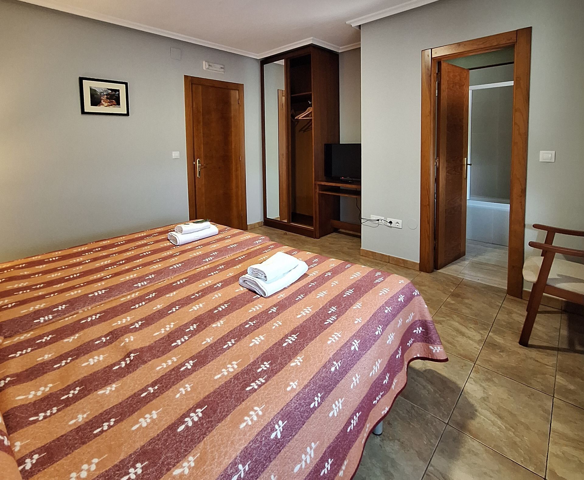 Quarto de hotel com cama, televisão, guarda-roupa e uma cadeira.