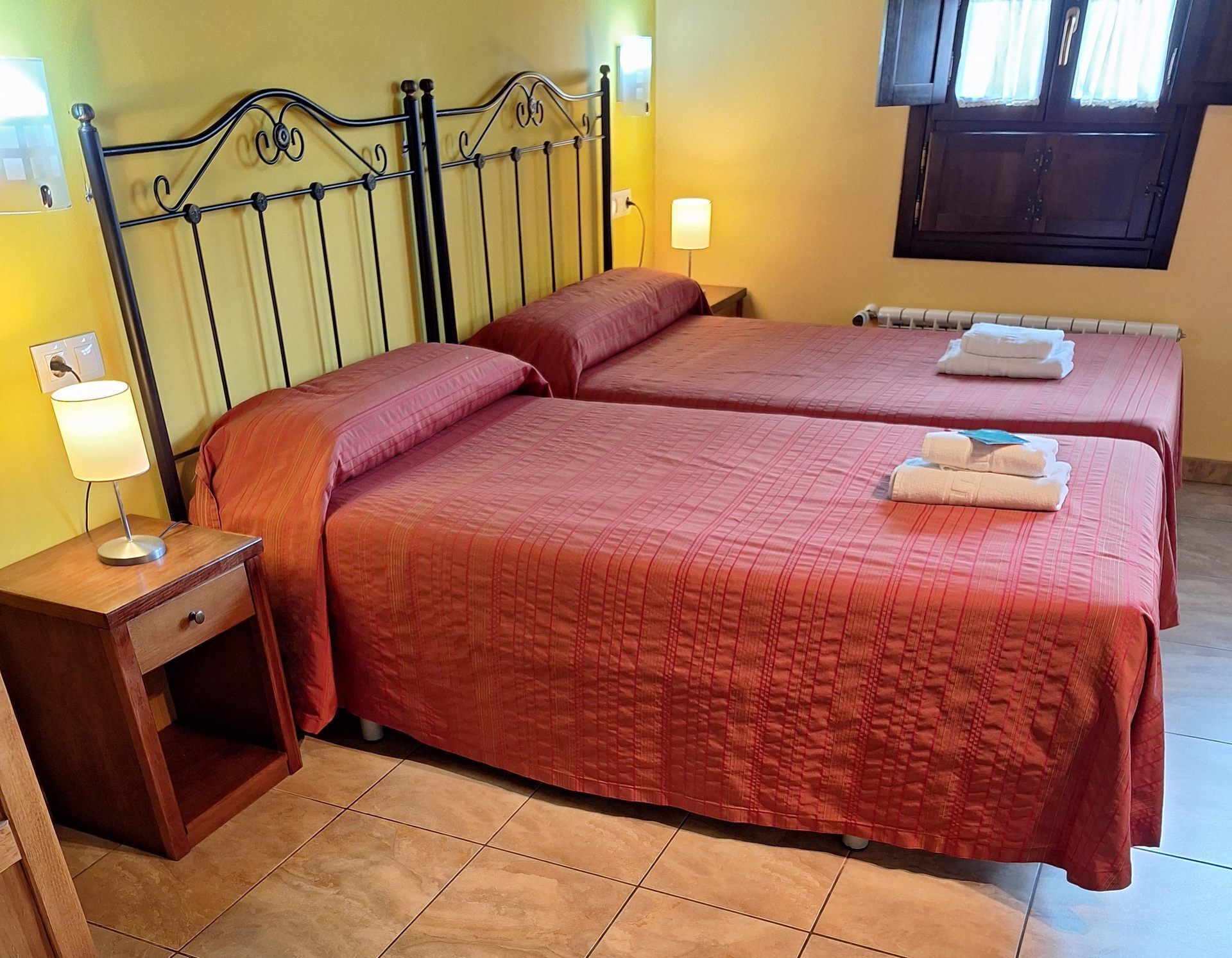 Quarto de hotel com duas camas, uma mesinha de cabeceira com abajur e uma janela com cortinas.