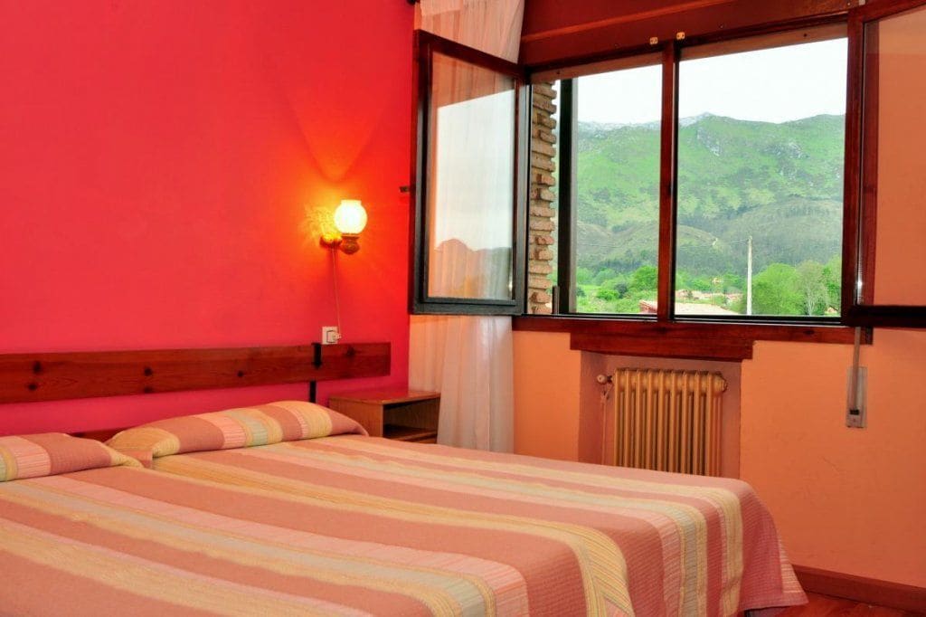 Un dormitorio con un ventanal grande, una cama con ropa de cama a rayas, una mesita de noche y una lámpara fijada a la pared.