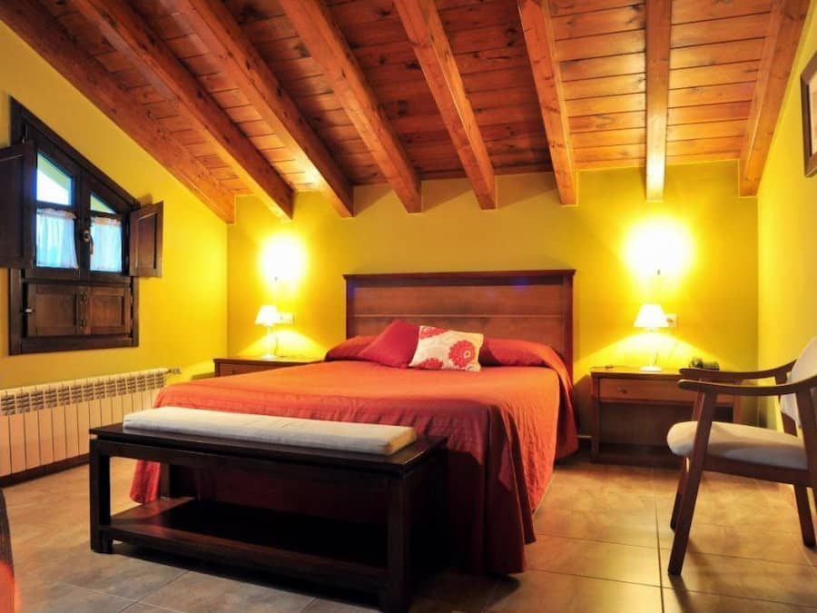 Een slaapkamer met een houten plafond, twee bedlampen, een bed met rode beddengoed, een nachtkastje, een stoel en een raam met gordijnen.