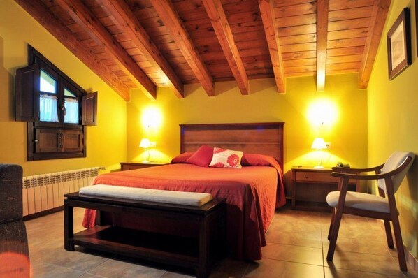 Un dormitorio con techo de madera, dos lámparas de noche, una cama con ropa de cama roja, una mesita de noche, una silla y una ventana con cortinas.