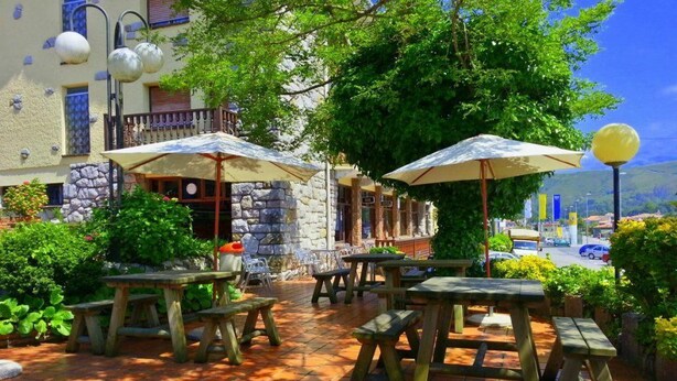 Een terras buiten met houten tafels en parasols, omgeven door groen en stenen muren.