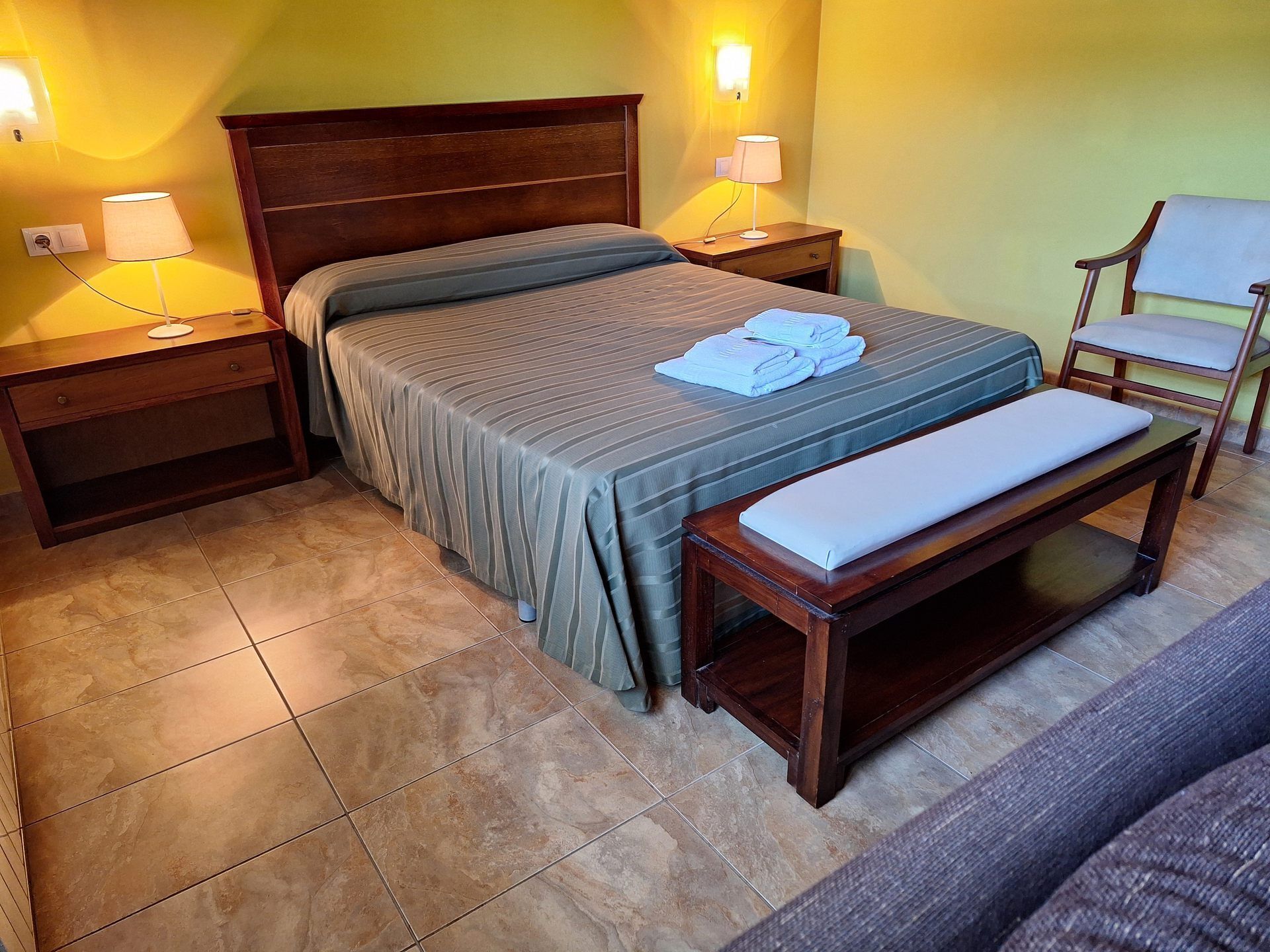 Habitación de hotel ordenada con cama, mesitas de noche, una silla y un banco.