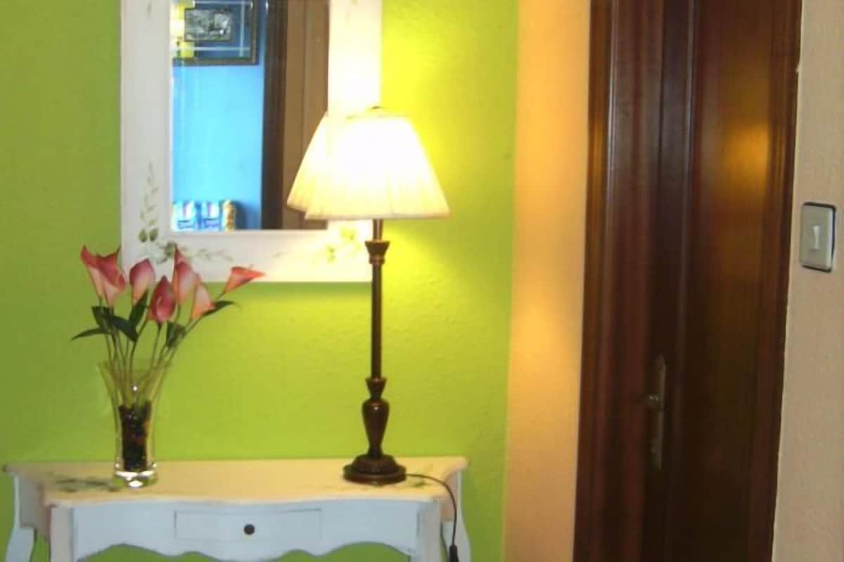 Um corredor com parede verde, mesa consola branca, espelho, luminária e um vaso com flores.