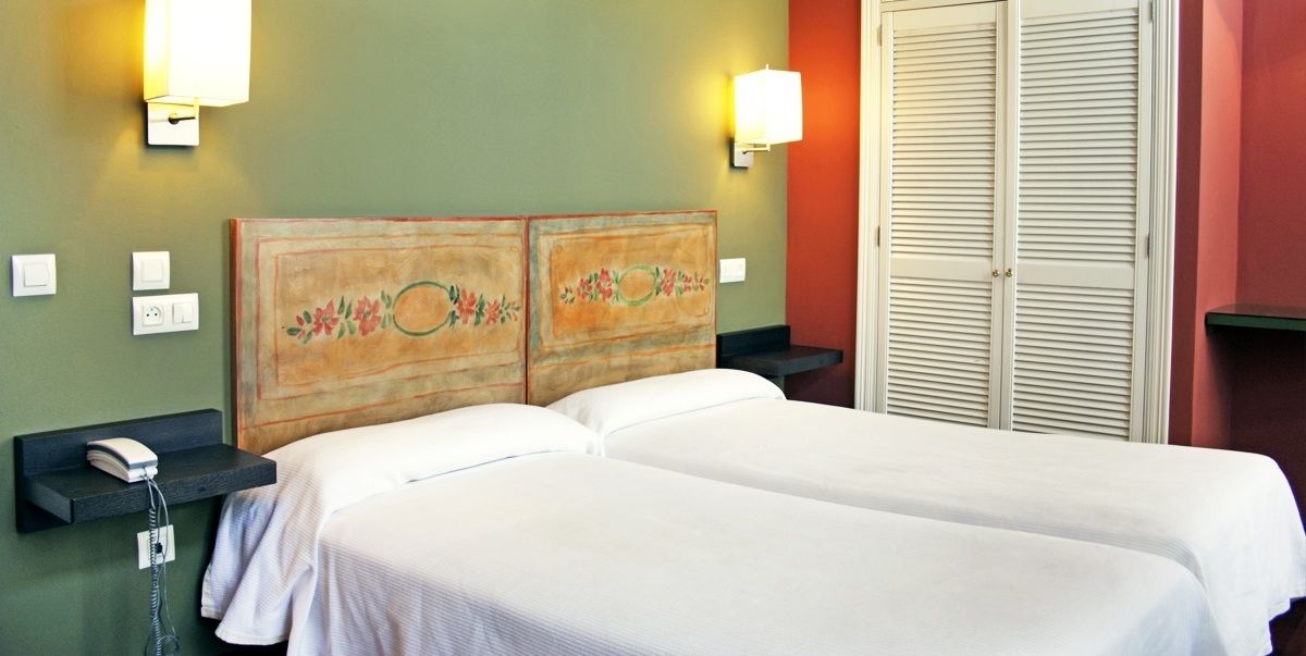 A hotel room with two beds, a wooden headboard with floral designs, bedside lamps, a phone on a nightstand, and a closet with sliding doors.