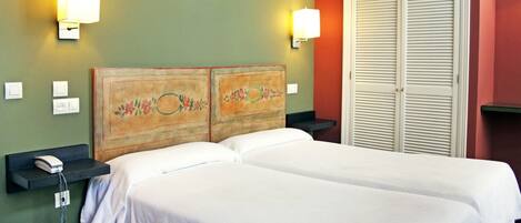 A hotel room with two beds, a wooden headboard with floral designs, bedside lamps, a phone on a nightstand, and a closet with sliding doors.