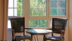 A room with two wicker chairs and a small round table, a large window with a view of greenery, and a curtain partially drawn. - La Colmena (Valdes)