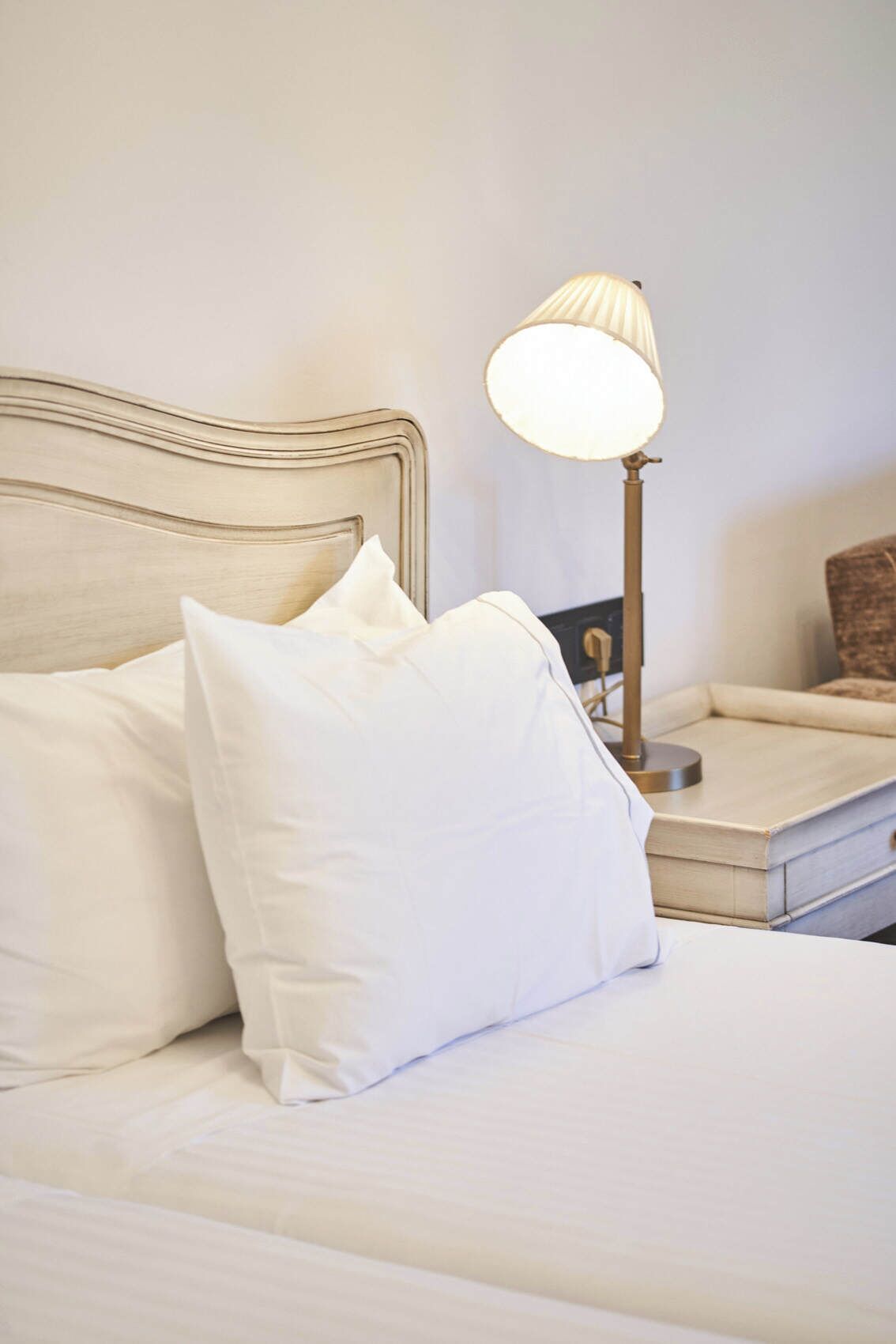 A neatly made bed with white linens and pillows, a bedside table with a lamp, and a small wooden nightstand.