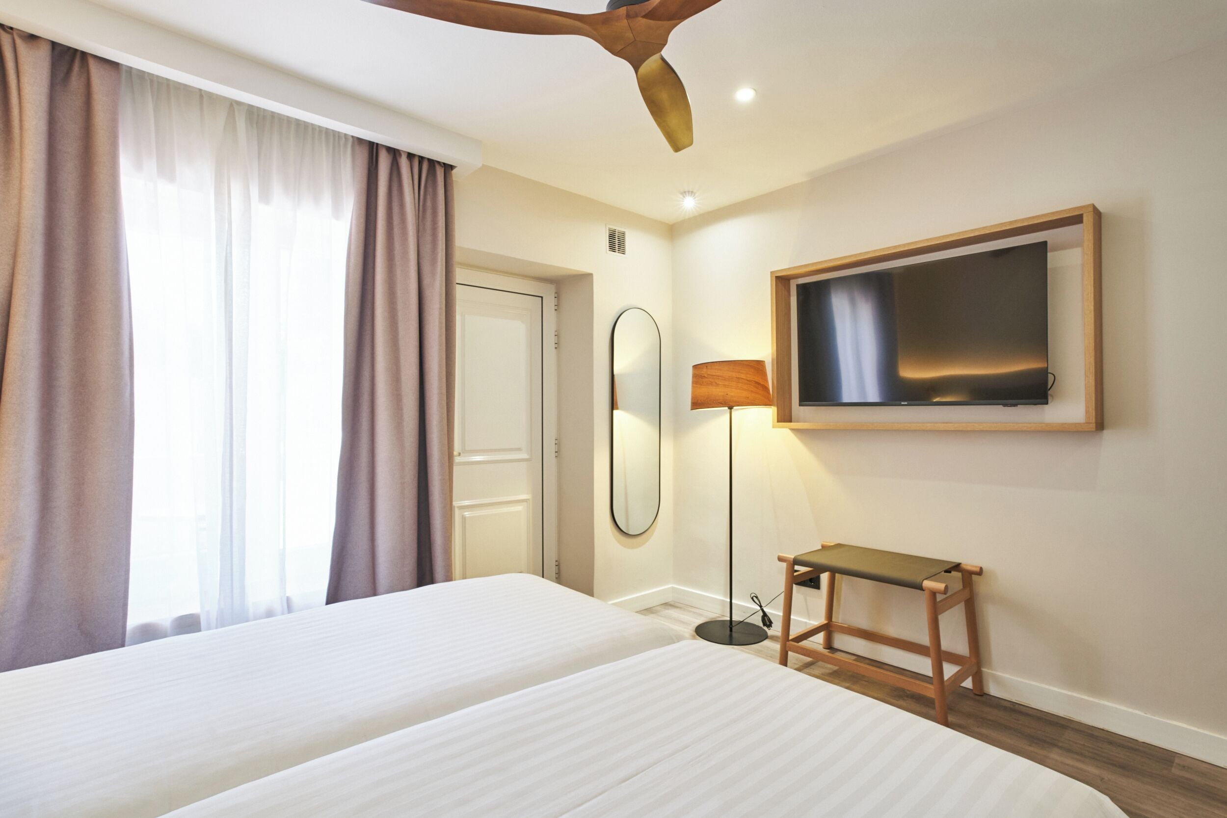 A hotel room with two beds, a ceiling fan, a TV mounted on the wall, a bedside lamp, and a small wooden table.