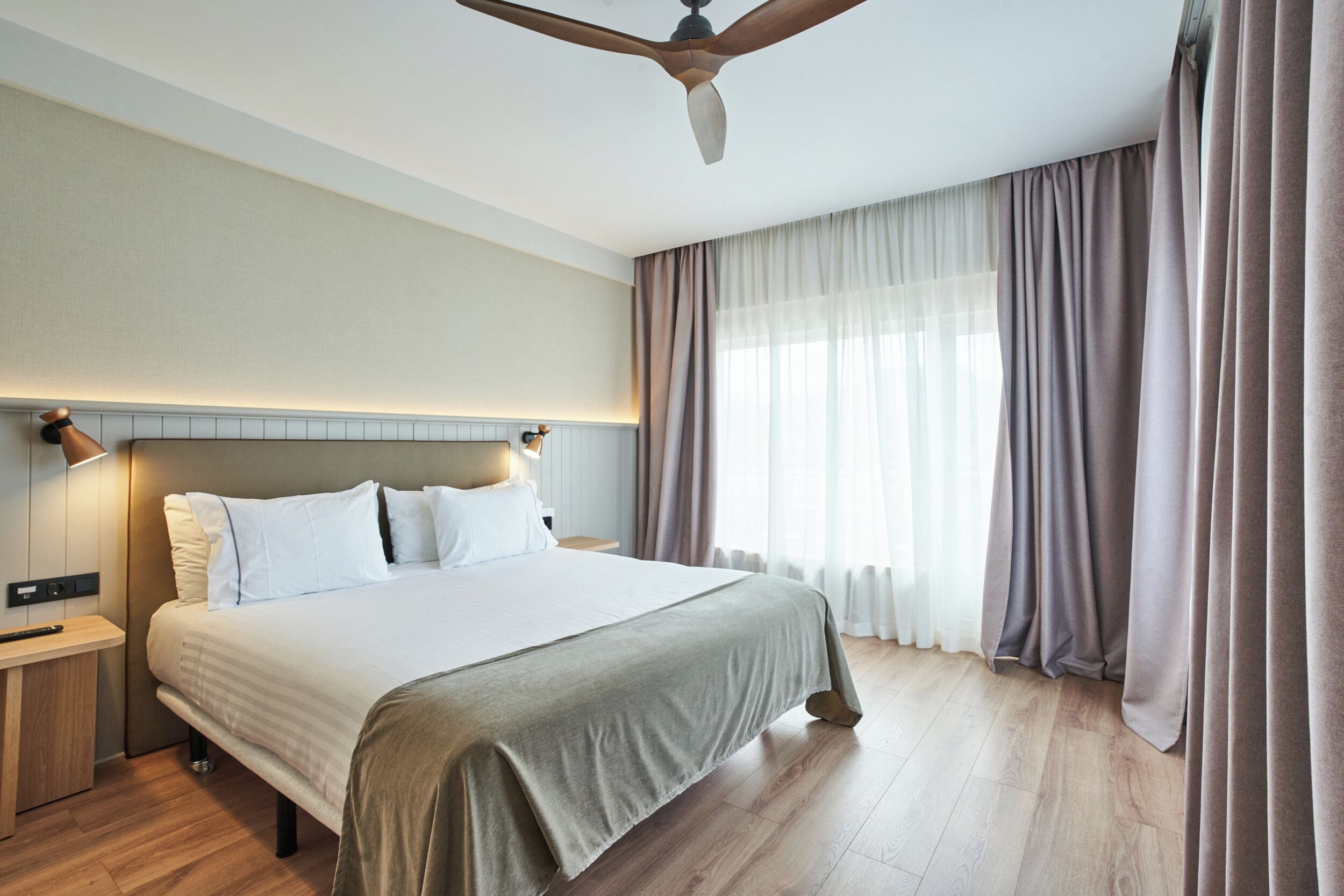 Habitación de hotel con una cama grande, piso de madera, ventilador de techo y ventana con cortinas.