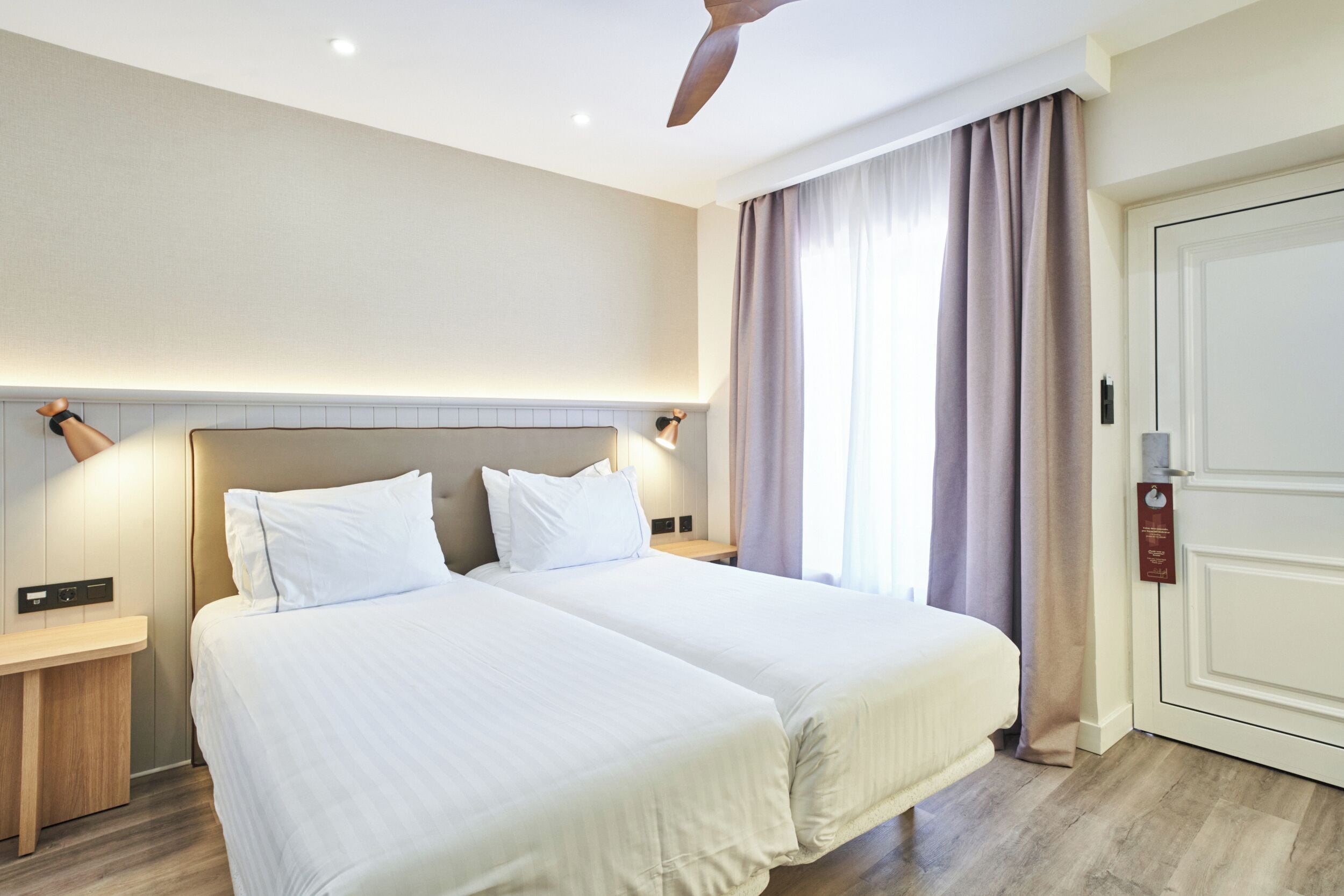 Una camera d'albergo con due letti, una ventola a soffitto, un comodino, una porta e una finestra con tende.
