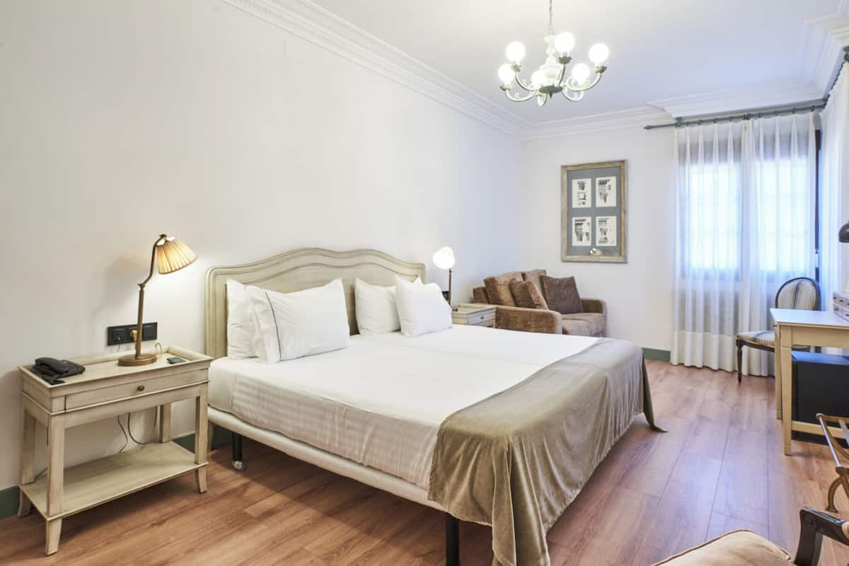 A hotel room with a bed, bedside table, lamp, chair, desk, and curtains.