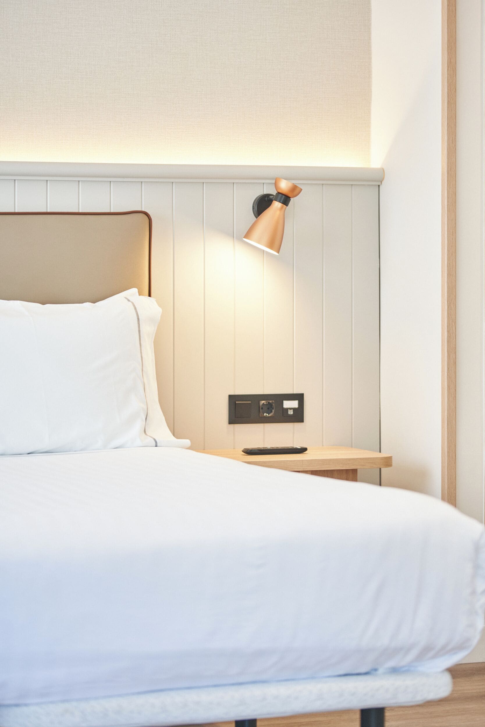A neatly made bed with a headboard, a bedside table with a lamp, and a wall-mounted control panel.