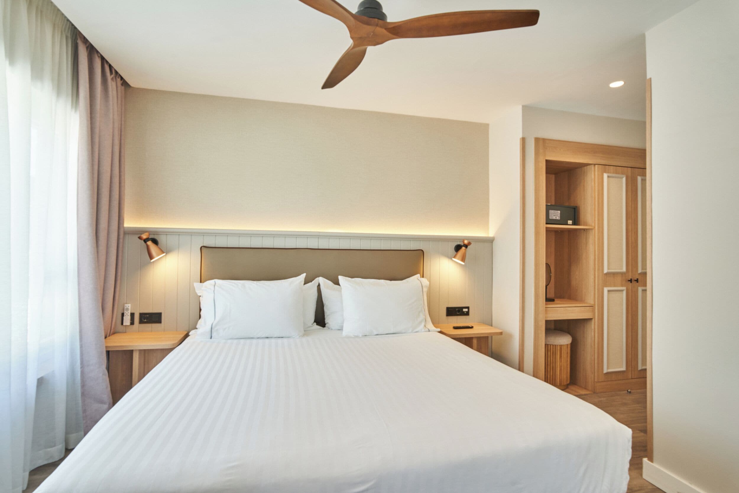 A hotel room with a large bed, wooden headboard, two bedside tables, a ceiling fan, and a wardrobe.