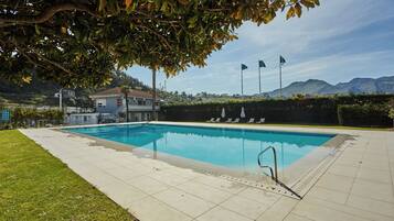 Una piscina rectangular con solárium empedrado, rodeada de césped y un seto. Hay banderas en postes al fondo, y un edificio con techo rojo es visible a lo lejos.