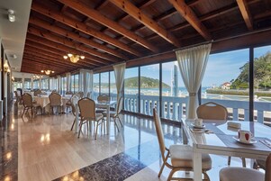 Uma ampla sala de jantar com vigas de madeira, janelas grandes e vista para o mar.