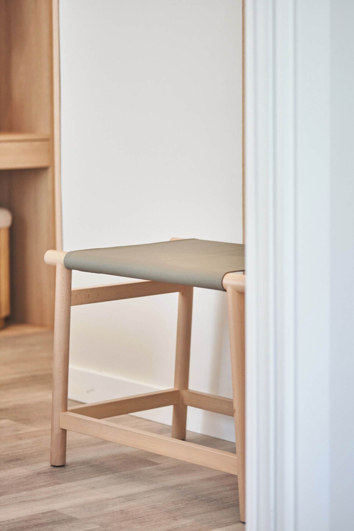 A wooden chair with a light-colored seat and backrest, positioned against a white wall and wooden floor.