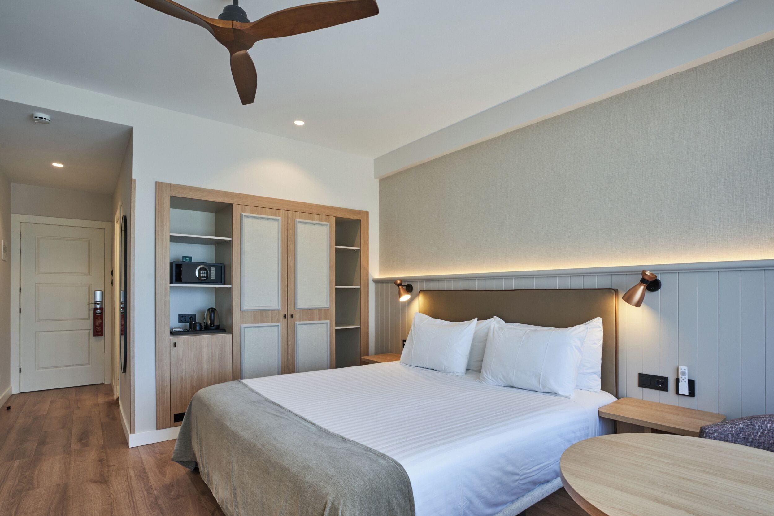 A hotel room with a bed, bedside table, built-in wardrobe, ceiling fan, and a wall-mounted light fixture.