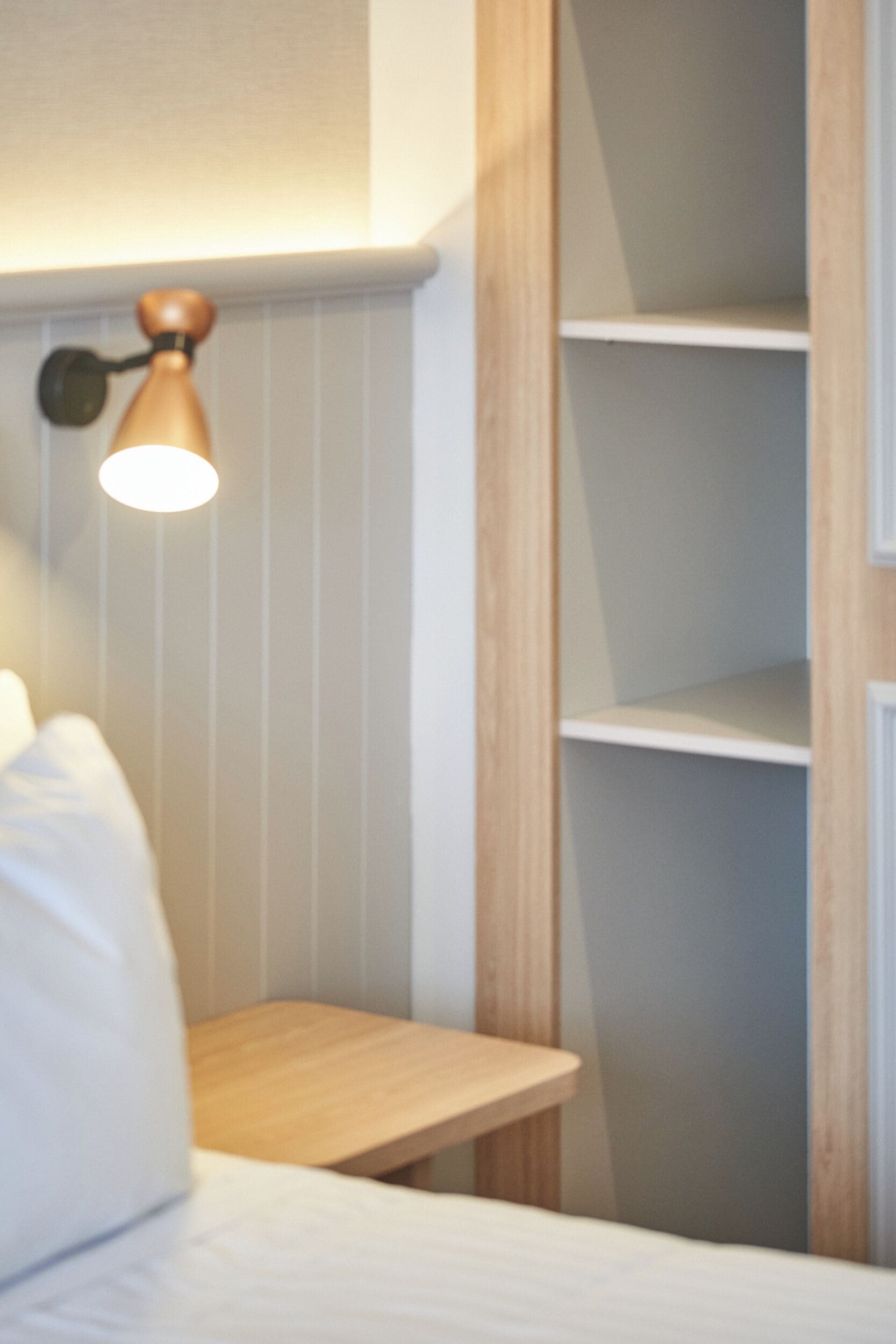 A room with a bed, a bedside table, a wall-mounted lamp, and a wooden shelf unit.