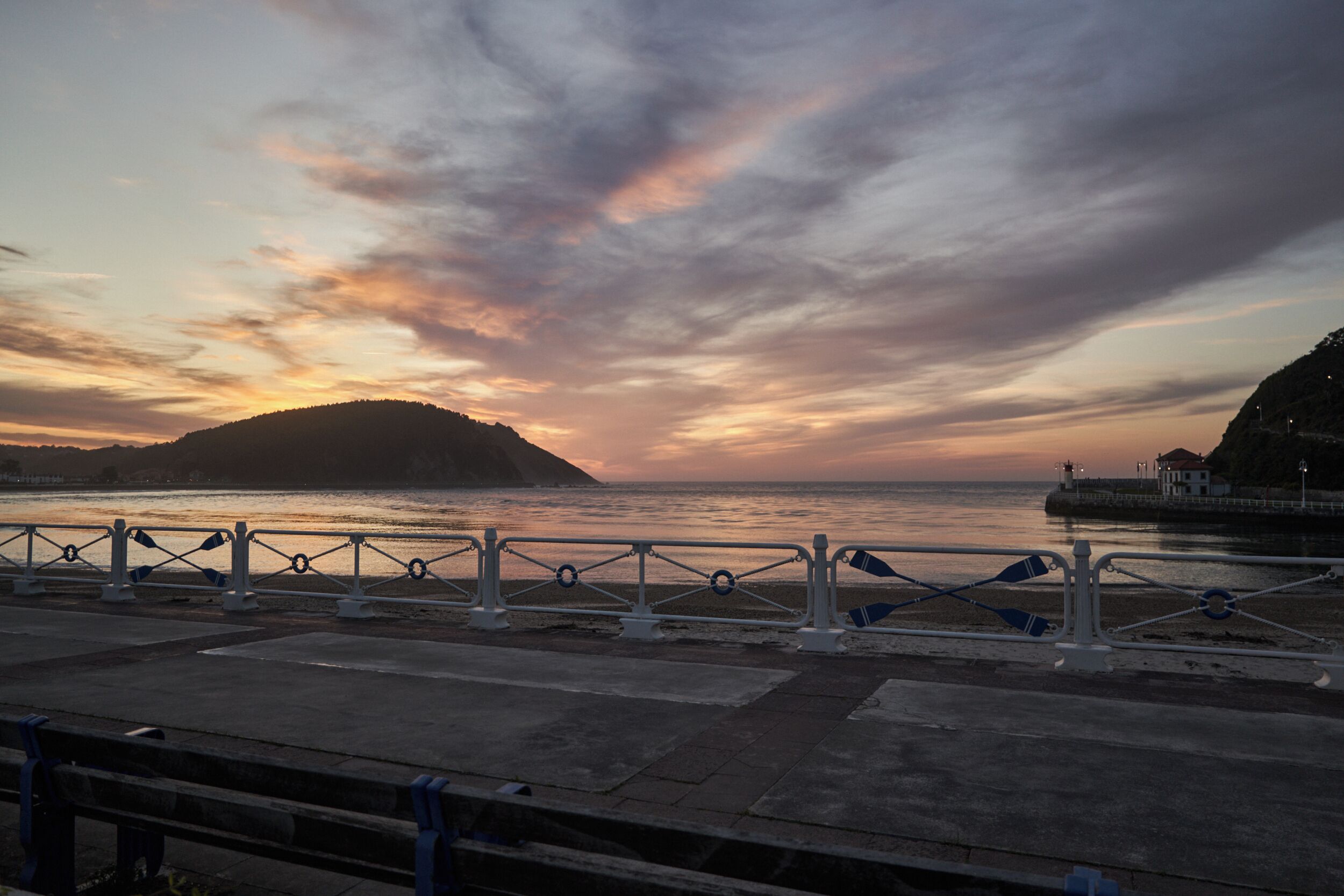 Um calçadão à beira-mar com guarda-corpo de metal, uma colina ao longe e um pôr do sol sobre a água.