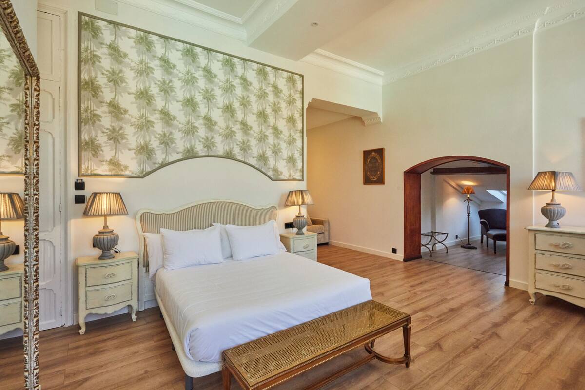 A bedroom with a large bed, two bedside tables with lamps, a wooden bench, a patterned wallpaper, and a view into another room.