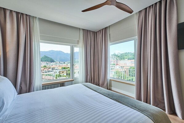 Una habitación de hotel con cama, ventilador de techo, ventana con cortinas y vistas a un paisaje urbano.
