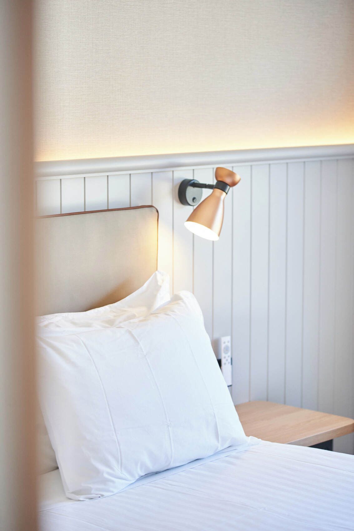 A neatly made bed with white linens, a wall-mounted lamp, and a wooden nightstand.