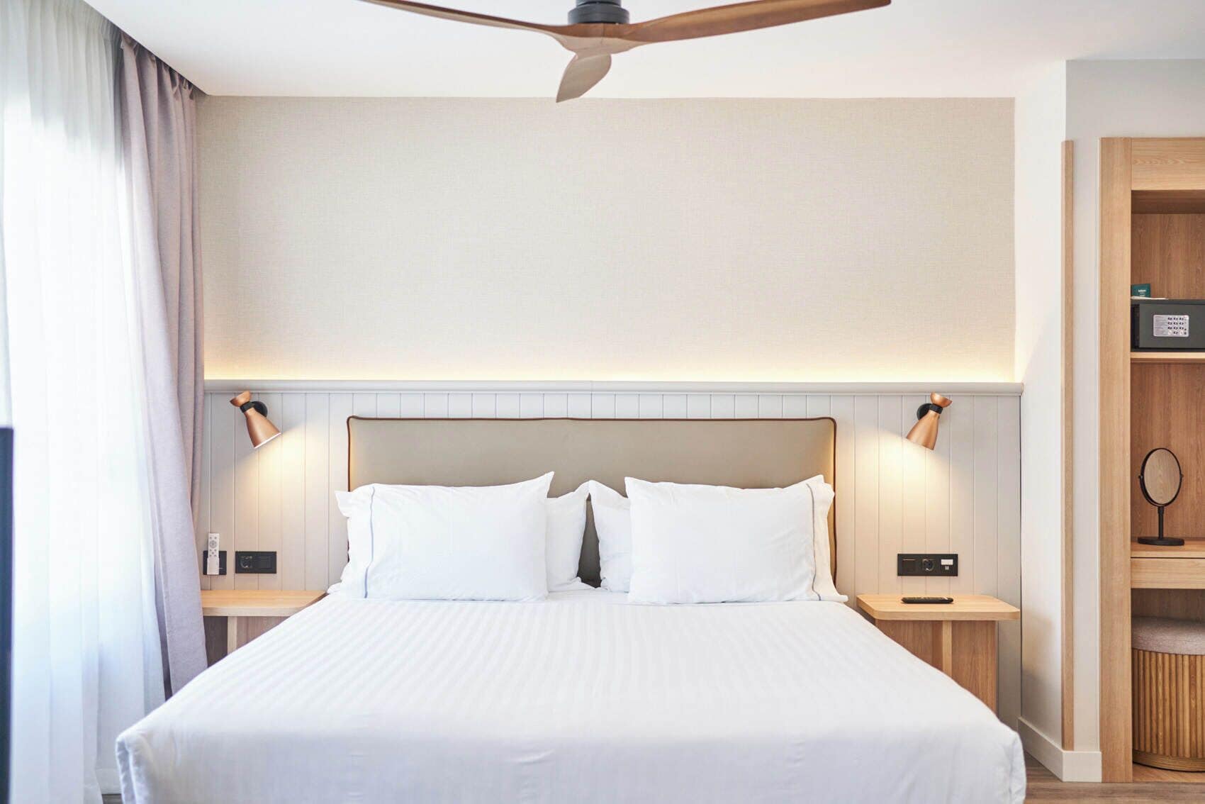 Habitación de hotel con una cama grande, lámparas de noche, ventilador de techo y un aparador de madera con espejo.